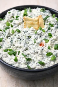 Black bowl of creamy spinach artichoke dip garnished with green onions and crackers.