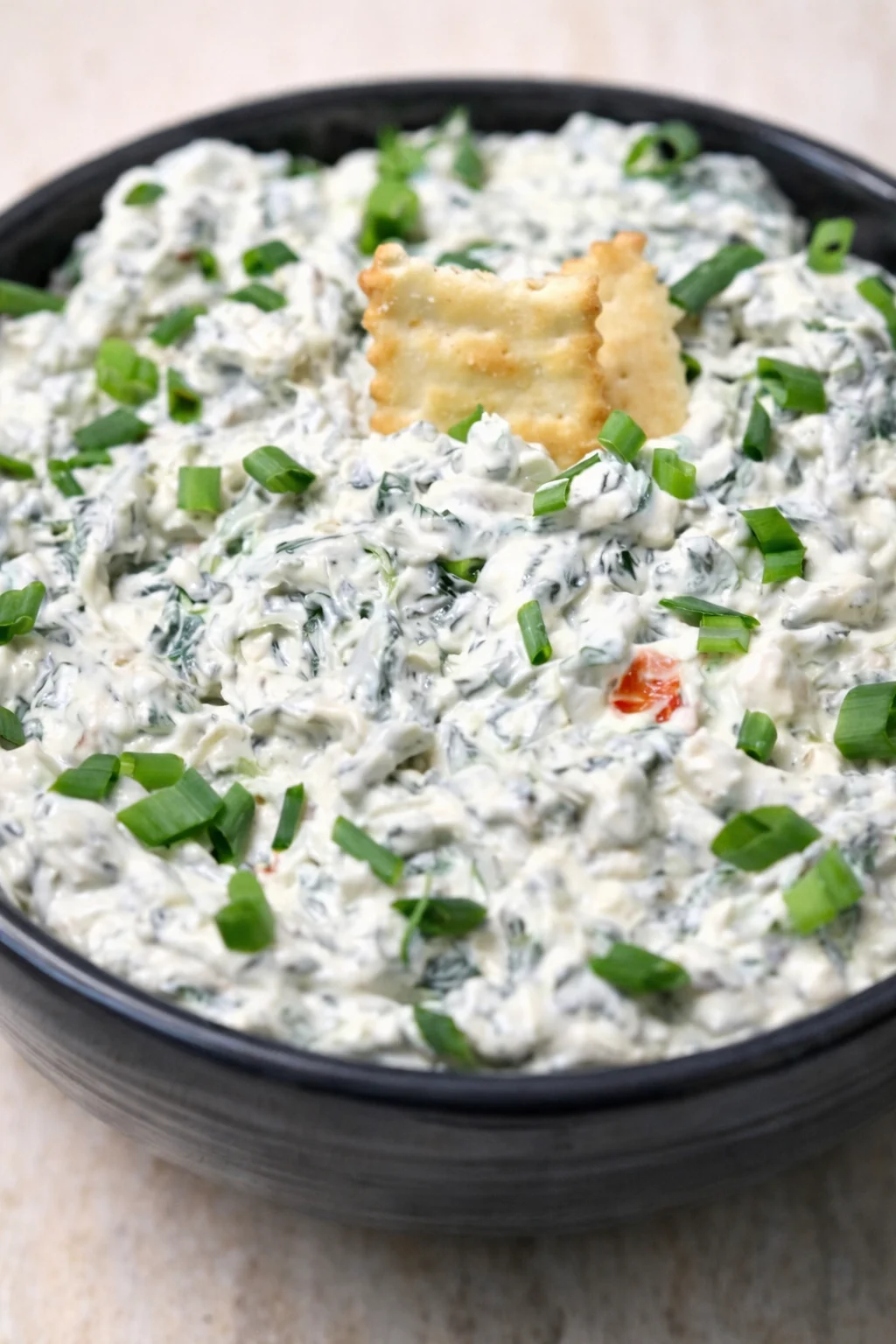 Black bowl of creamy spinach artichoke dip garnished with green onions and crackers.