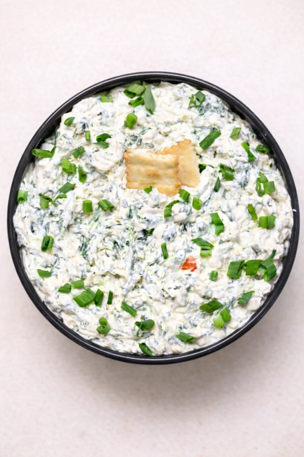 Top-down bowl of creamy spinach dip topped with chopped green onions and crackers.