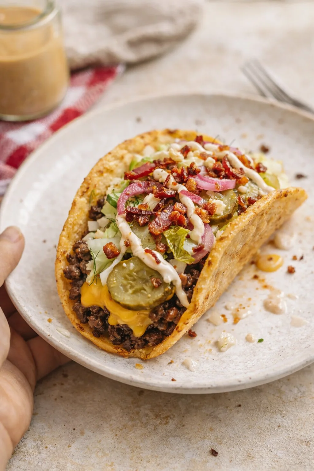 A loaded crispy beef taco on a speckled plate, topped with bacon, pickles, onions, herbs, and a creamy white drizzle.