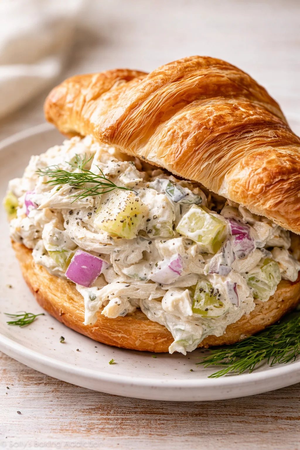 Golden-brown croissant sandwich filled with creamy chicken salad and dill garnish on a plate.