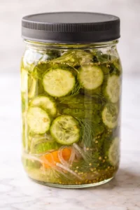 A glass jar filled with sliced cucumbers, dill, onions, carrot, and mustard seeds in brine.