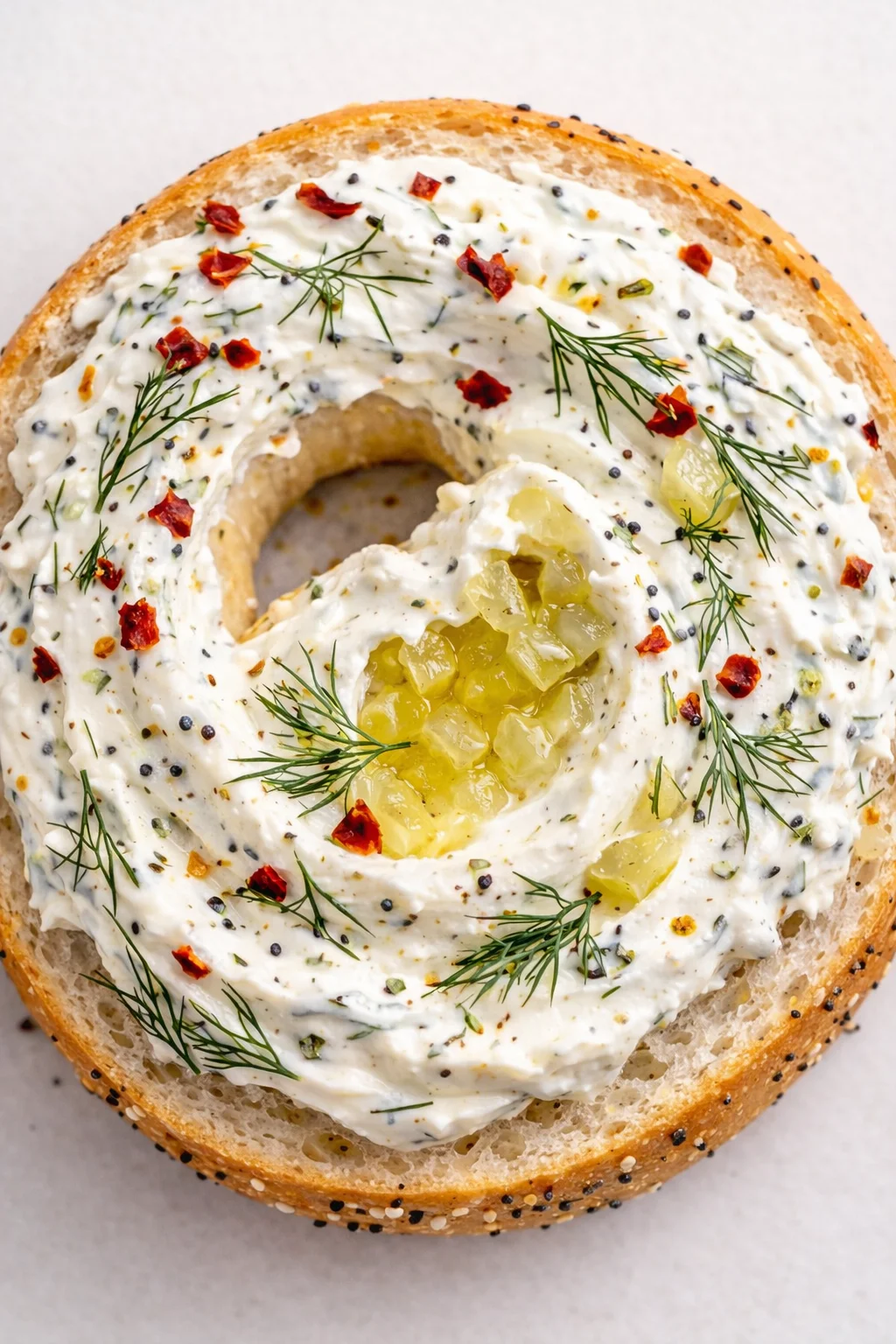 top-down view of an everything bagel with herbed cream cheese, dill, chili flakes, and diced yellow pickles