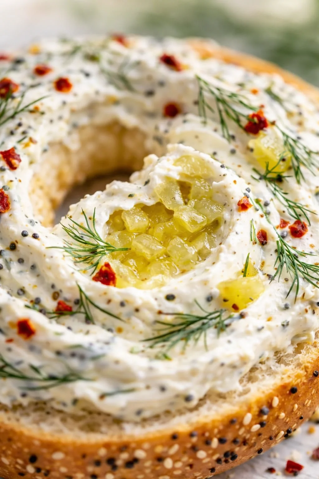 Close-up of an everything bagel topped with herb cream cheese, dill sprigs, red pepper flakes, and pineapple chunks in the center.