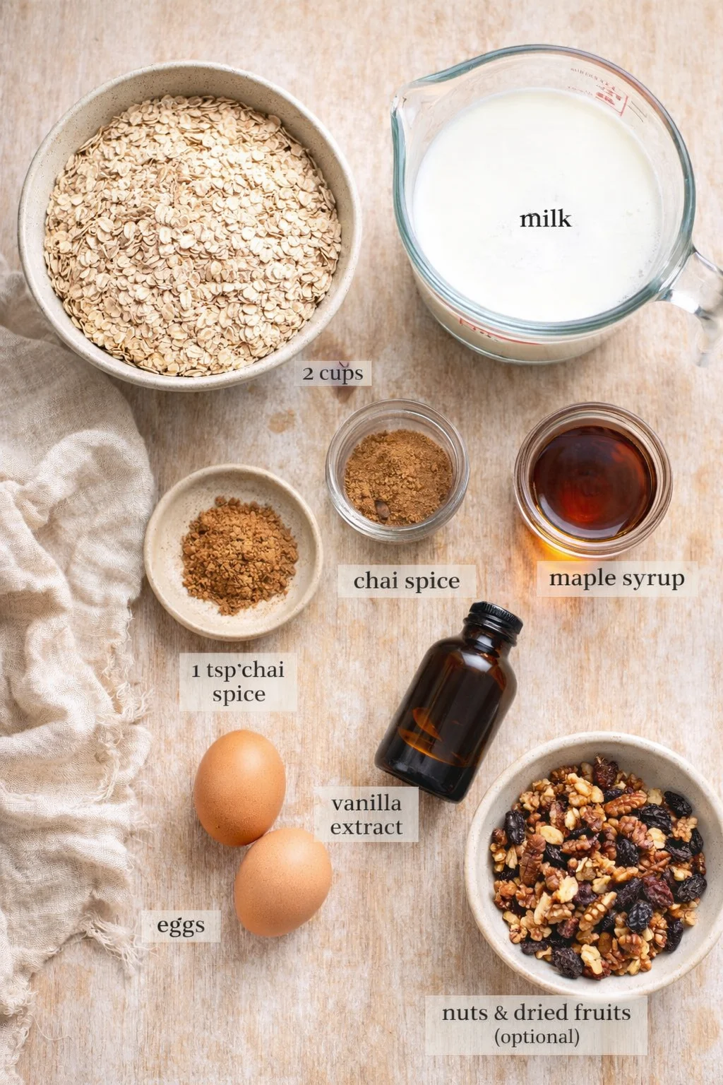Top-down view of oats, milk, spices, maple syrup, eggs, vanilla, nuts, and dried fruits arranged on a wooden surface.