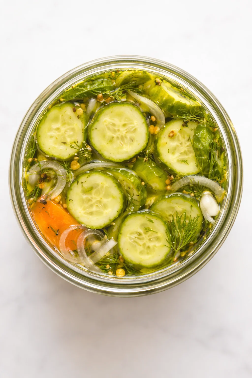 Top-down view of a glass jar filled with cucumber slices, dill, onions, carrot, garlic, and brine.