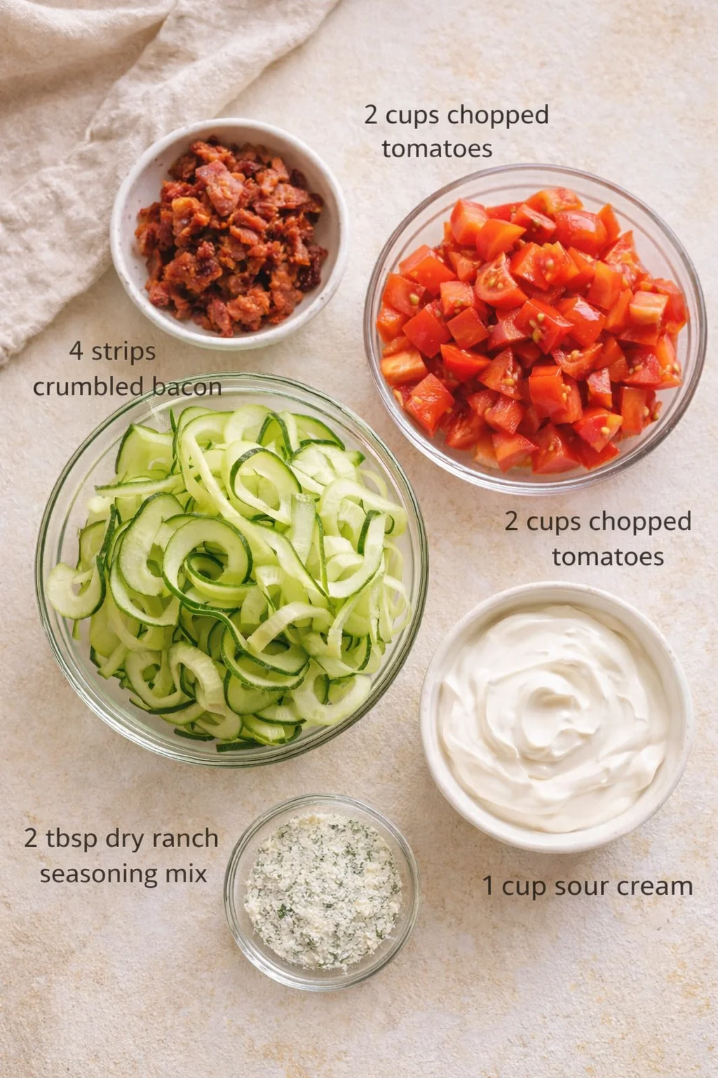 Top-down view of bowls with chopped tomatoes, cucumber ribbons, bacon bits, sour cream, and ranch seasoning.