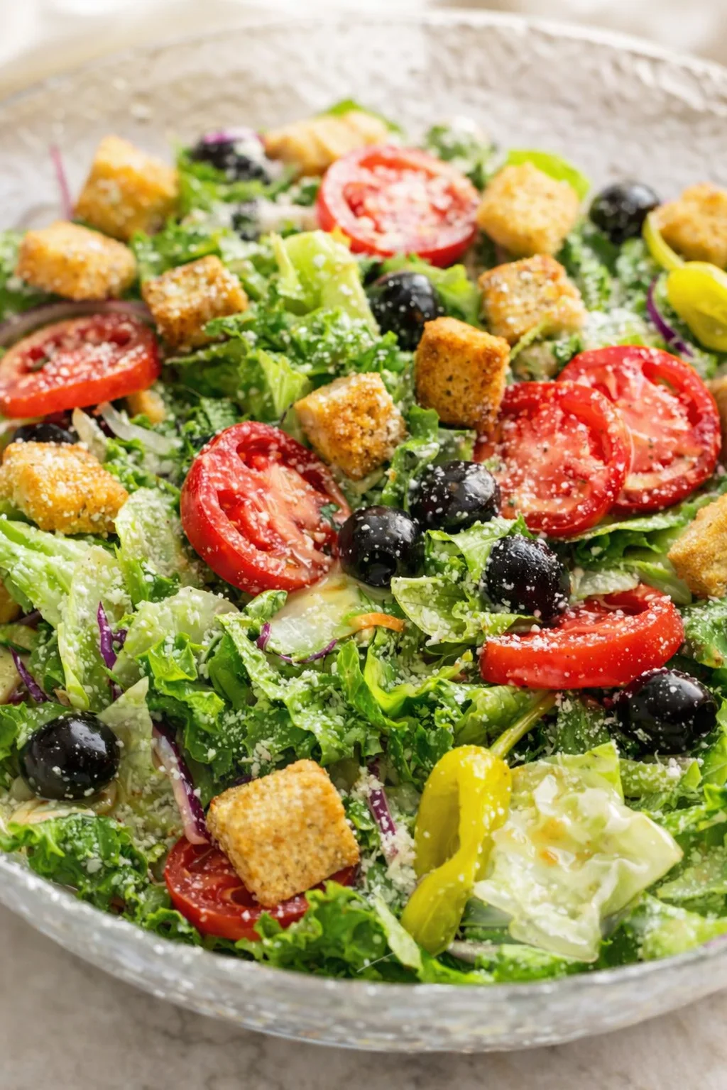 Colorful mixed-greens salad with tomato slices, black olives, croutons, and parmesan in a speckled bowl.