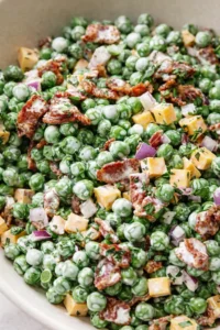 Close-up of green peas tossed with cheddar cheese cubes, bacon, red onion, and herbs in a creamy dressing