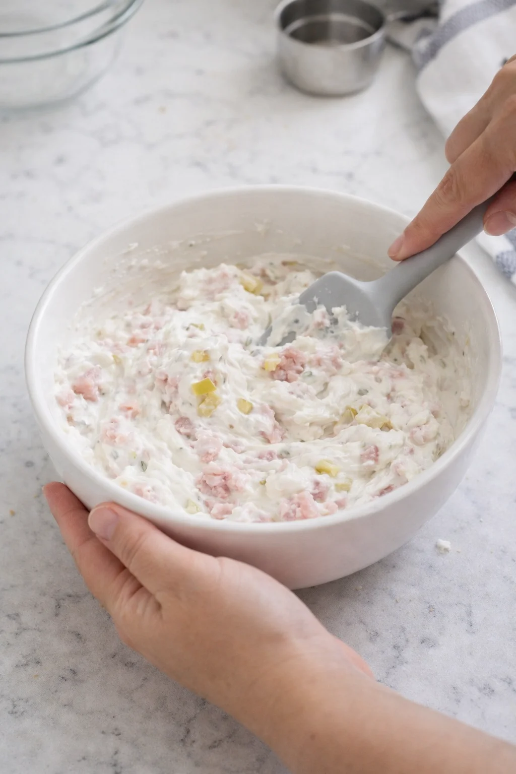 A person stirs a creamy salad with pink and yellow diced ingredients in a white bowl.