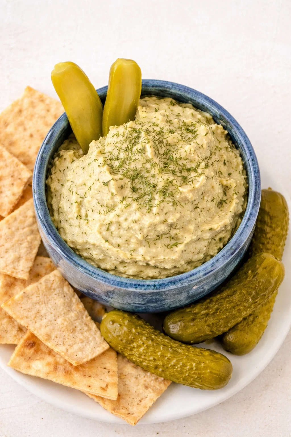 Blue bowl of creamy hummus topped with dill, surrounded by pita crackers and pickles.