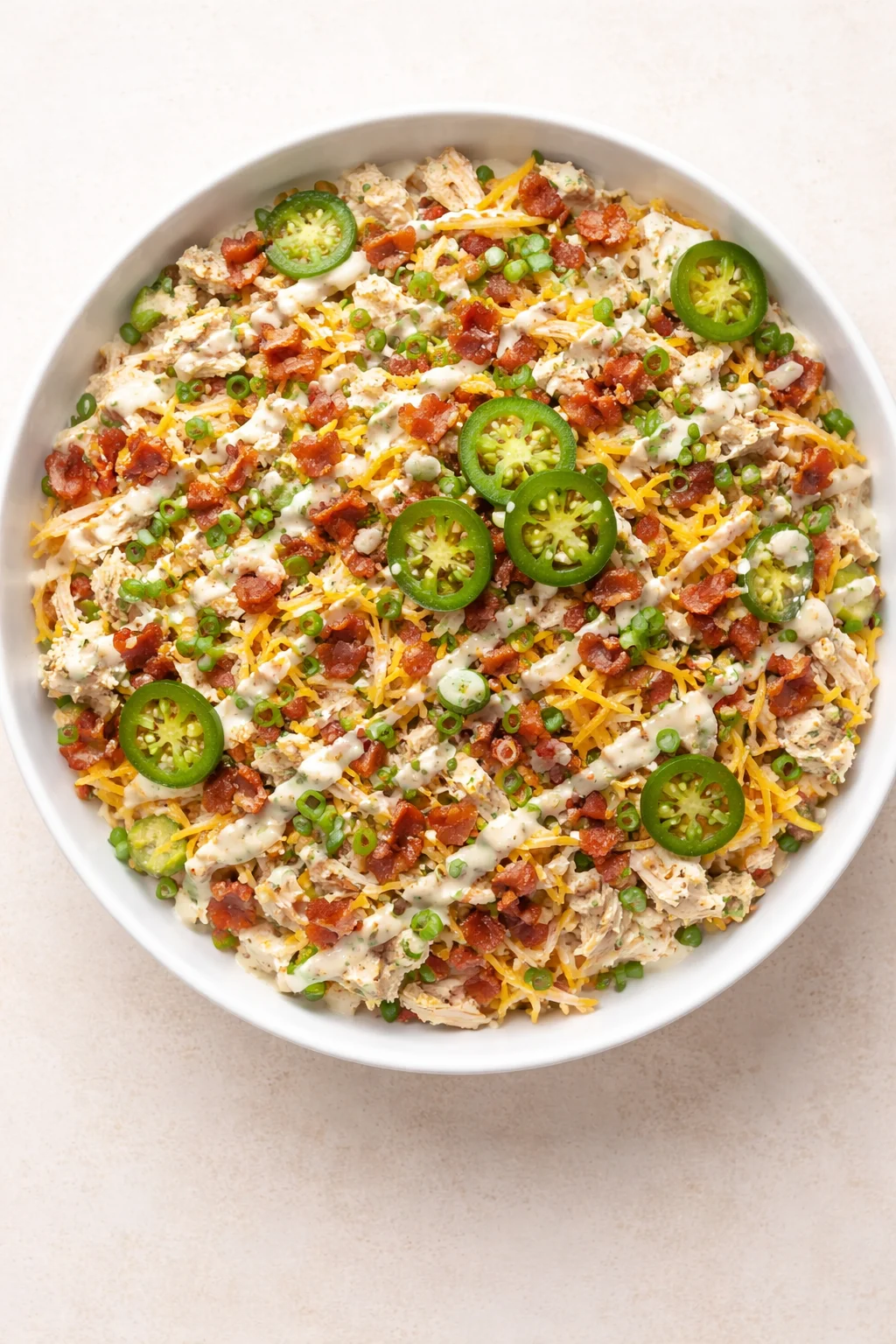 round white bowl of creamy chicken dip topped with bacon bits, jalapeño slices, and green onions.