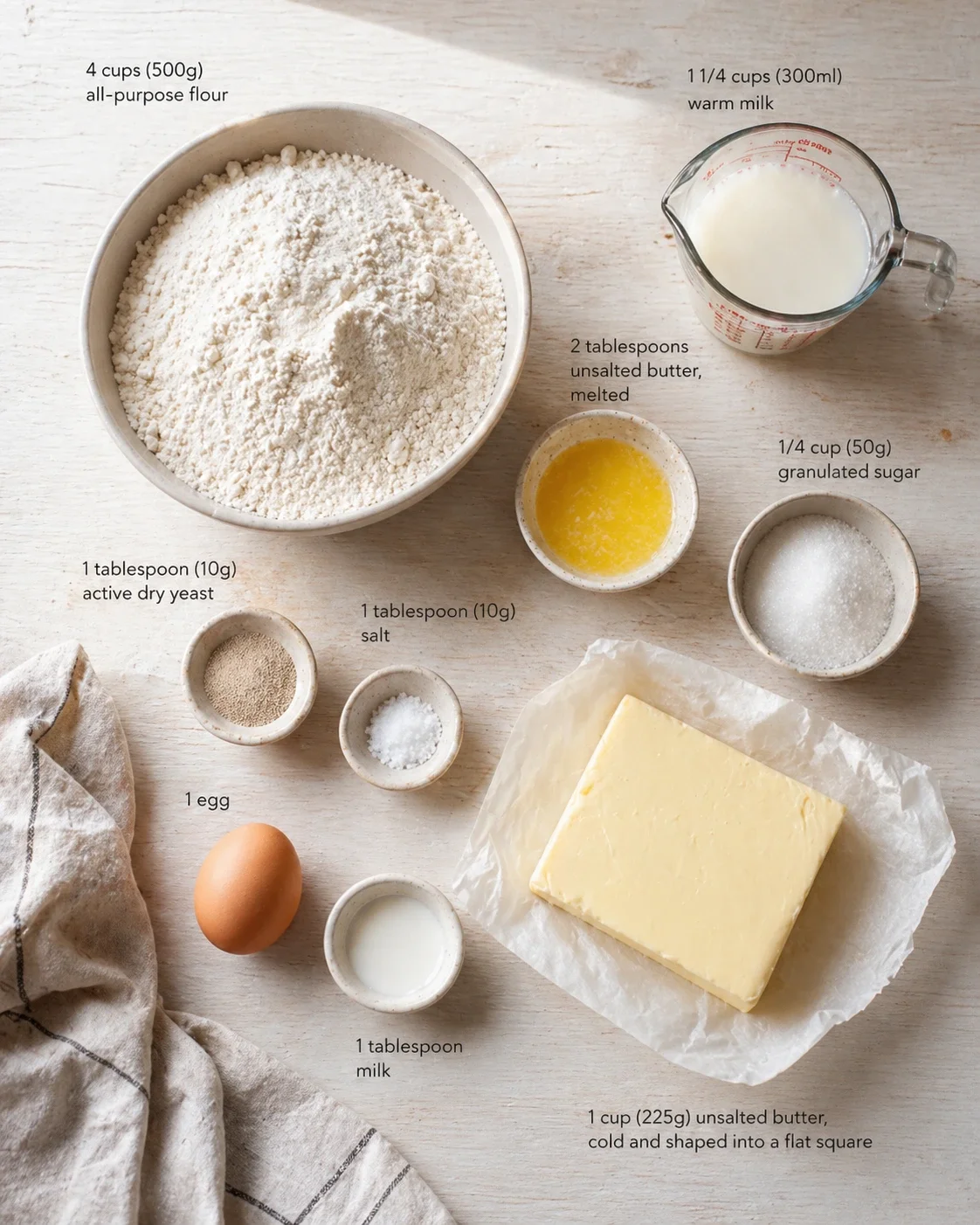 Top-down view of baking ingredients laid out on a pale wooden surface: flour, milk, butter, egg, sugar, and yeast.