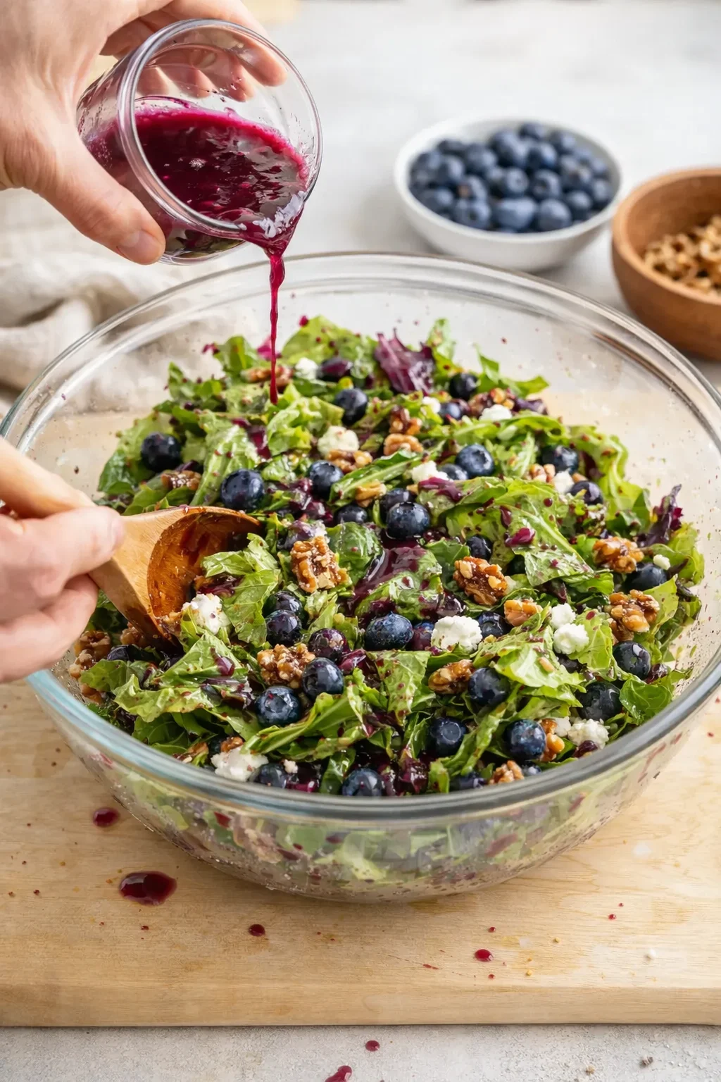 Hands pouring purple beet dressing into a glass bowl of mixed greens, blueberries, feta, and pecans.