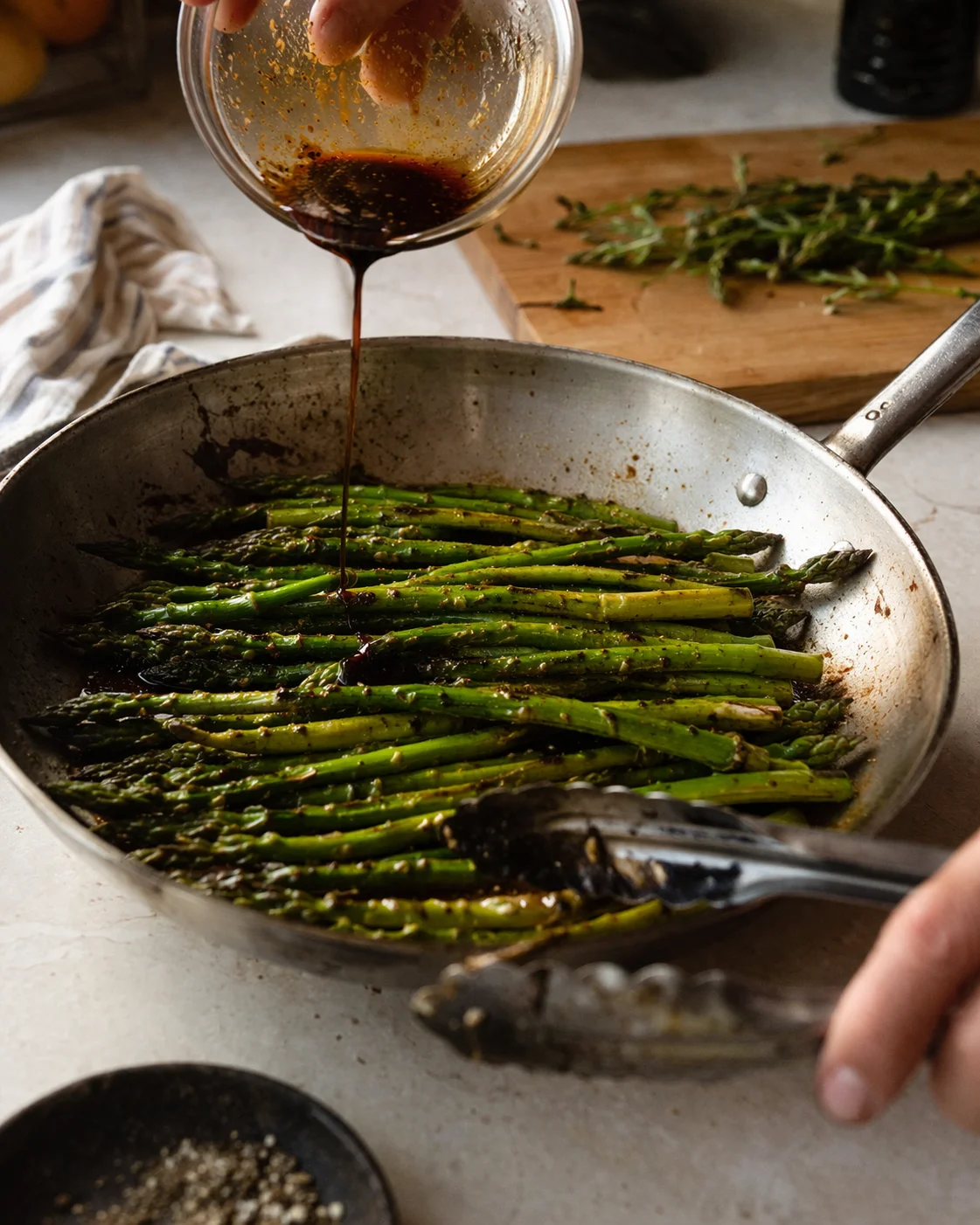 Green asparagus in a skillet while a dark glaze pours from a glass bowl onto the spears.