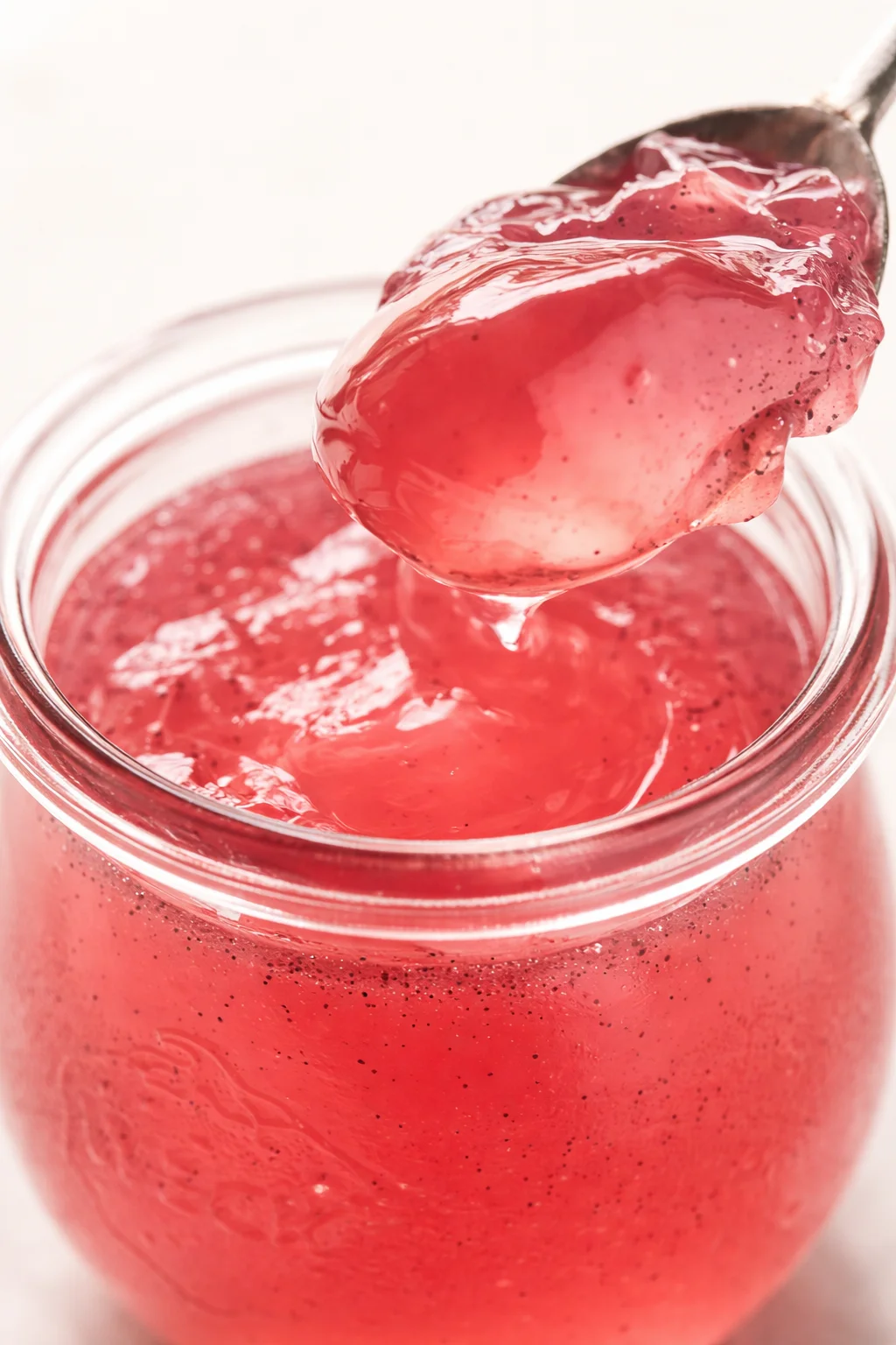 Close-up of a glass jar filled with glossy pink jam, with a spoon lifting jam from the jar