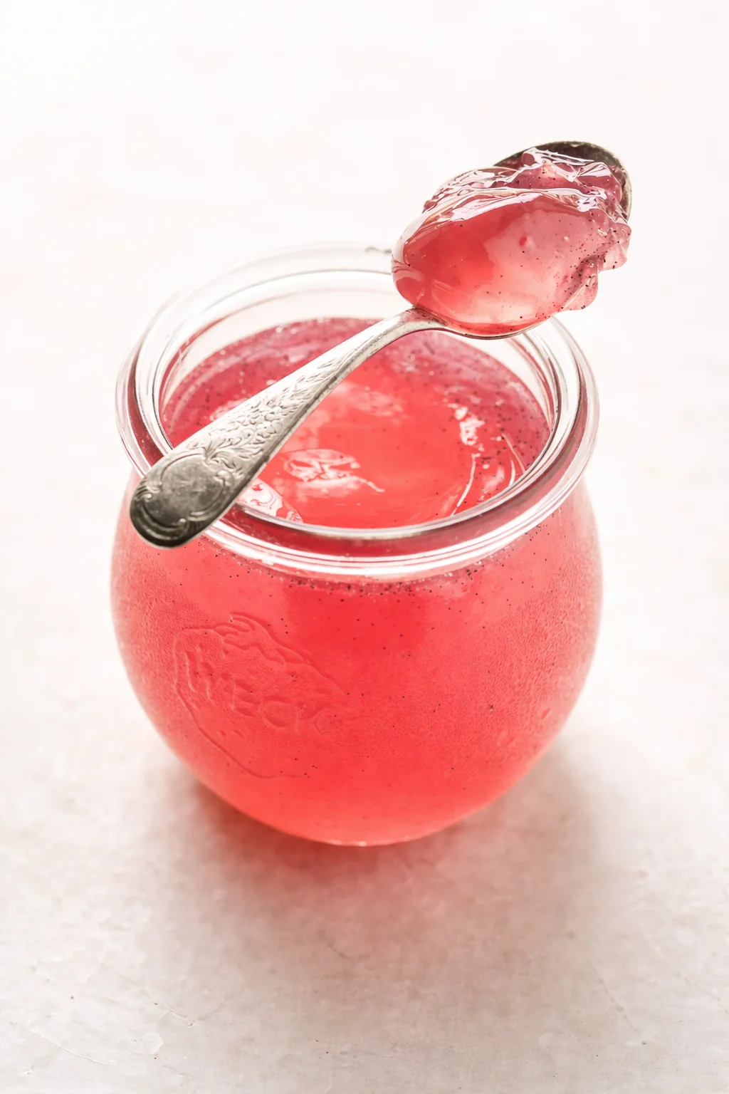 Small glass jar of pink raspberry jam with an ornate spoon resting on the rim.