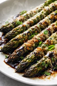 close-up of roasted asparagus spears topped with cheese, chopped herbs, and balsamic glaze on a white plate.