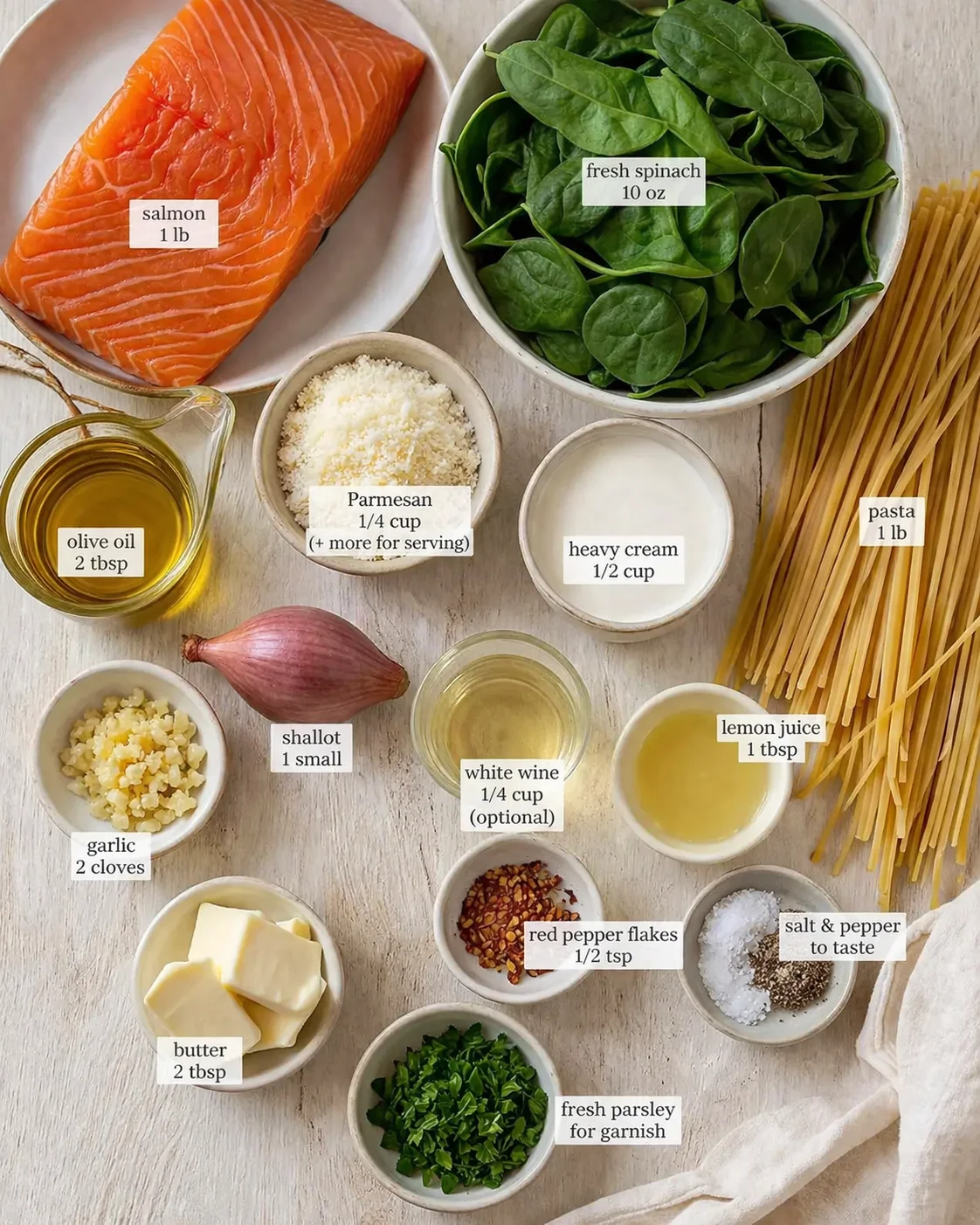 Assorted ingredients for salmon spinach pasta arranged on a light wood surface with labeled cards.