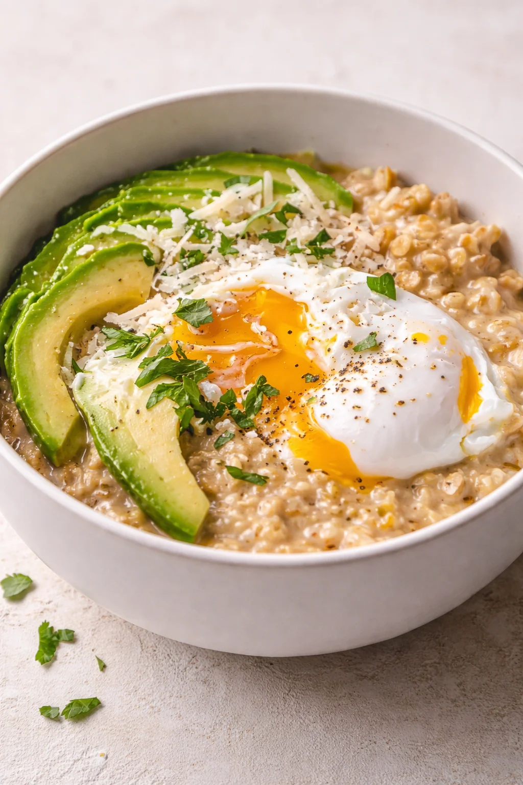 Creamy oatmeal with avocado slices, a runny egg, cheese, and herbs in a white bowl.