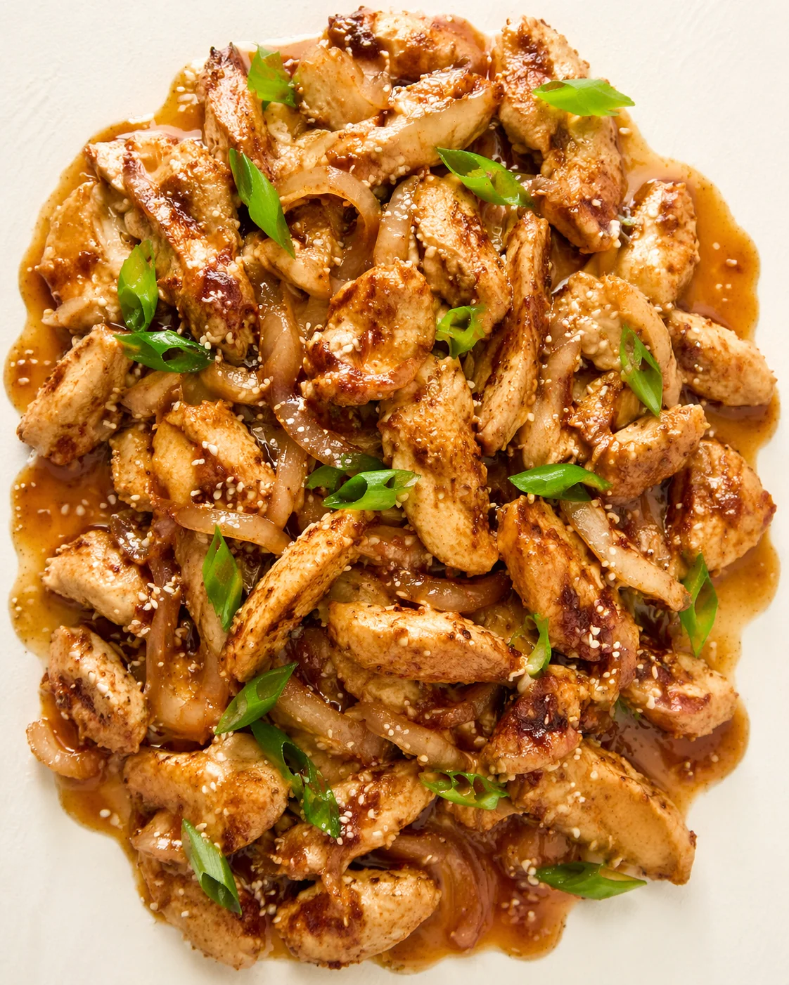 Plated sesame chicken with onions in a glossy brown sauce, sprinkled with sesame seeds and green onions.