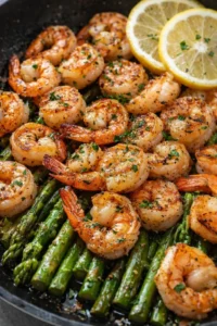 Plump sautéed shrimp atop green asparagus with lemon slices in a dark skillet.
