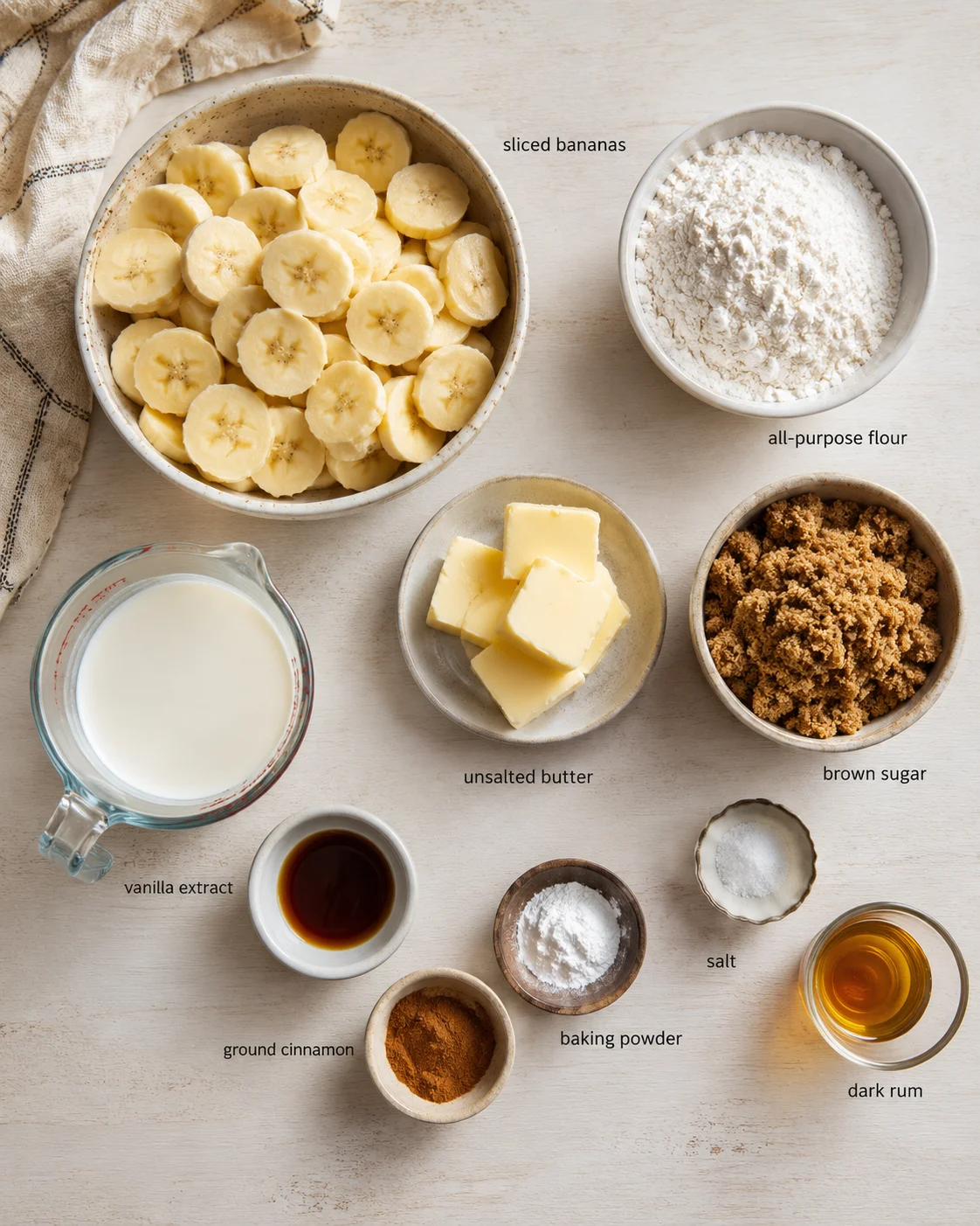 top-down flatlay of baking ingredients: sliced bananas, flour, butter, brown sugar, milk, rum, and spices
