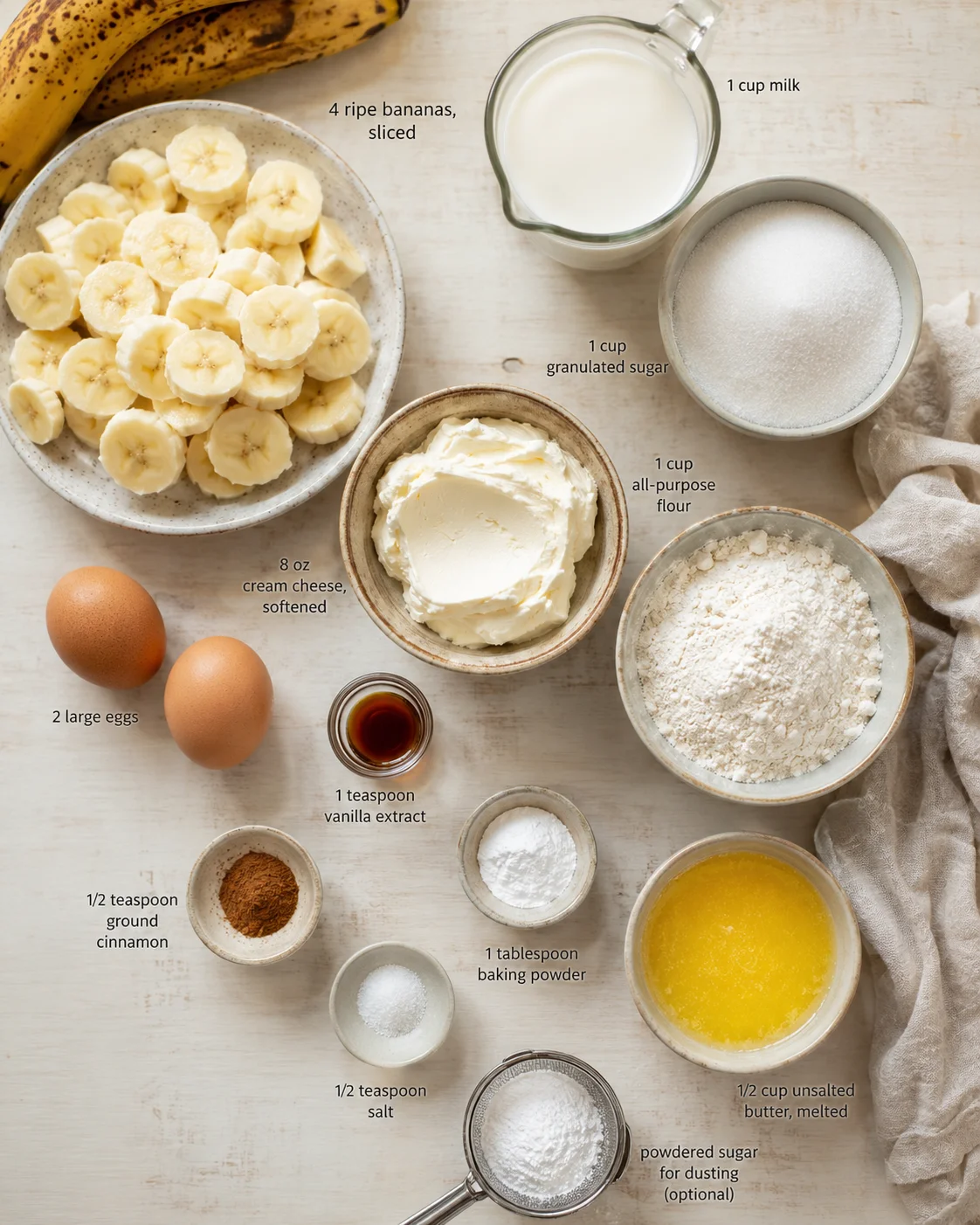 Flat lay of bananas, eggs, flour, sugar, cream cheese, melted butter, milk, and spices on a light wooden surface.