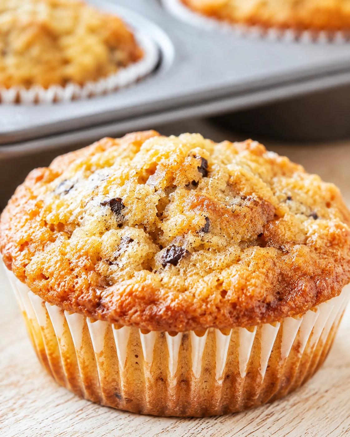 close-up of a golden streusel-topped banana muffin in a white liner on a wooden surface.