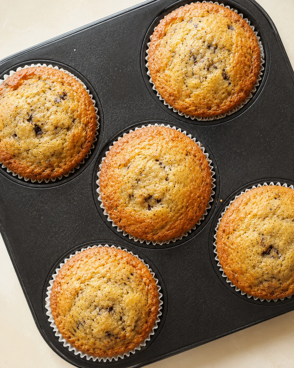 Golden blueberry muffins in white paper liners in a dark muffin tin on a light countertop.
