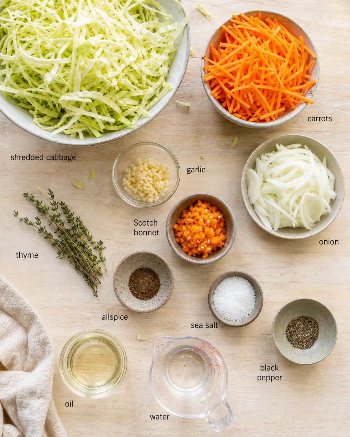 Overhead view of shredded cabbage, carrots, onions, garlic, thyme, and scotch bonnet pepper arranged in bowls.