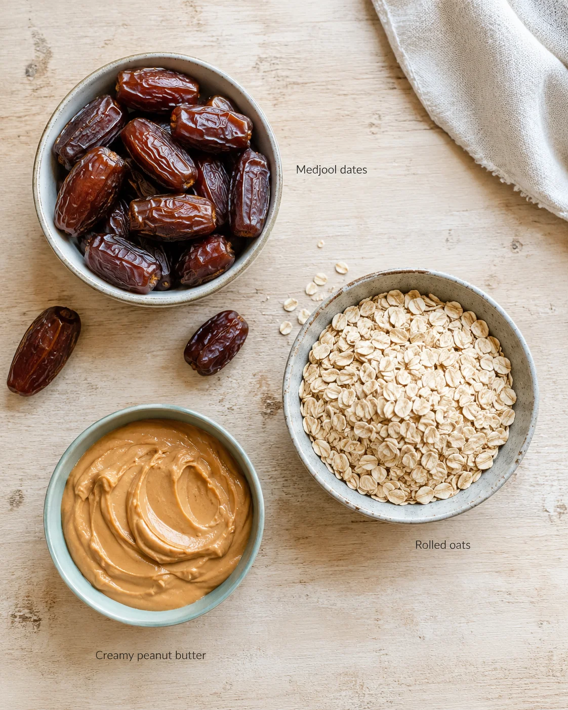 Top-down view of medjool dates, rolled oats, and creamy peanut butter arranged in bowls on a wooden surface.