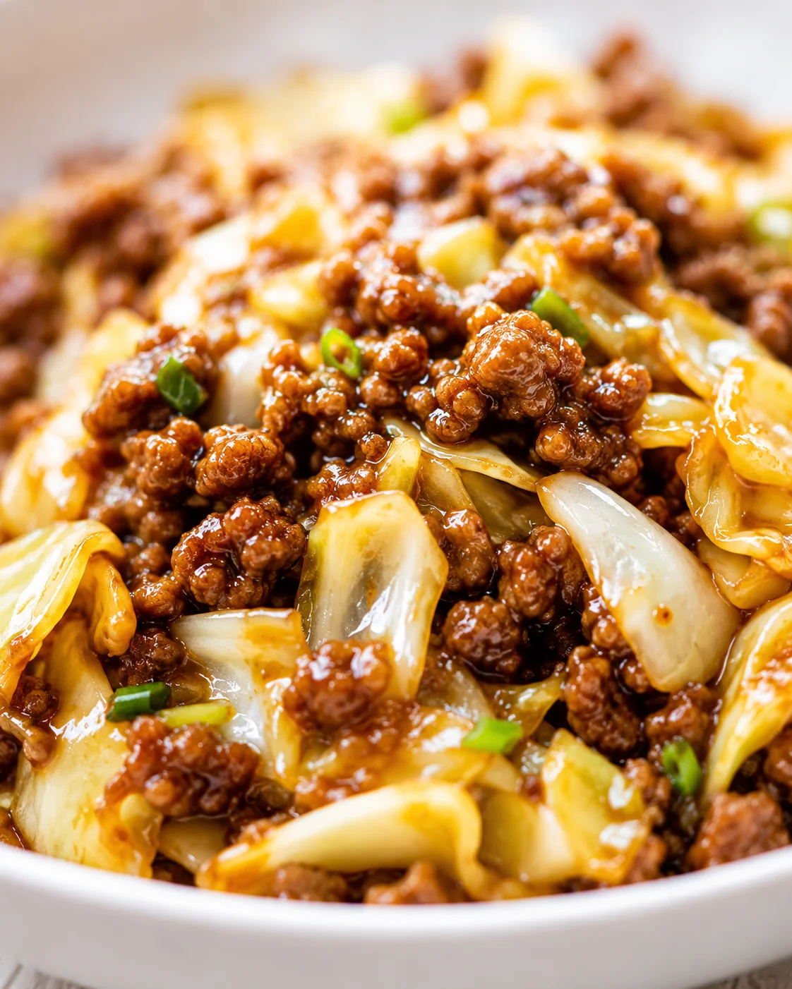 Close-up of glossy ground beef with onions in brown sauce, in a white bowl with green onion garnish.