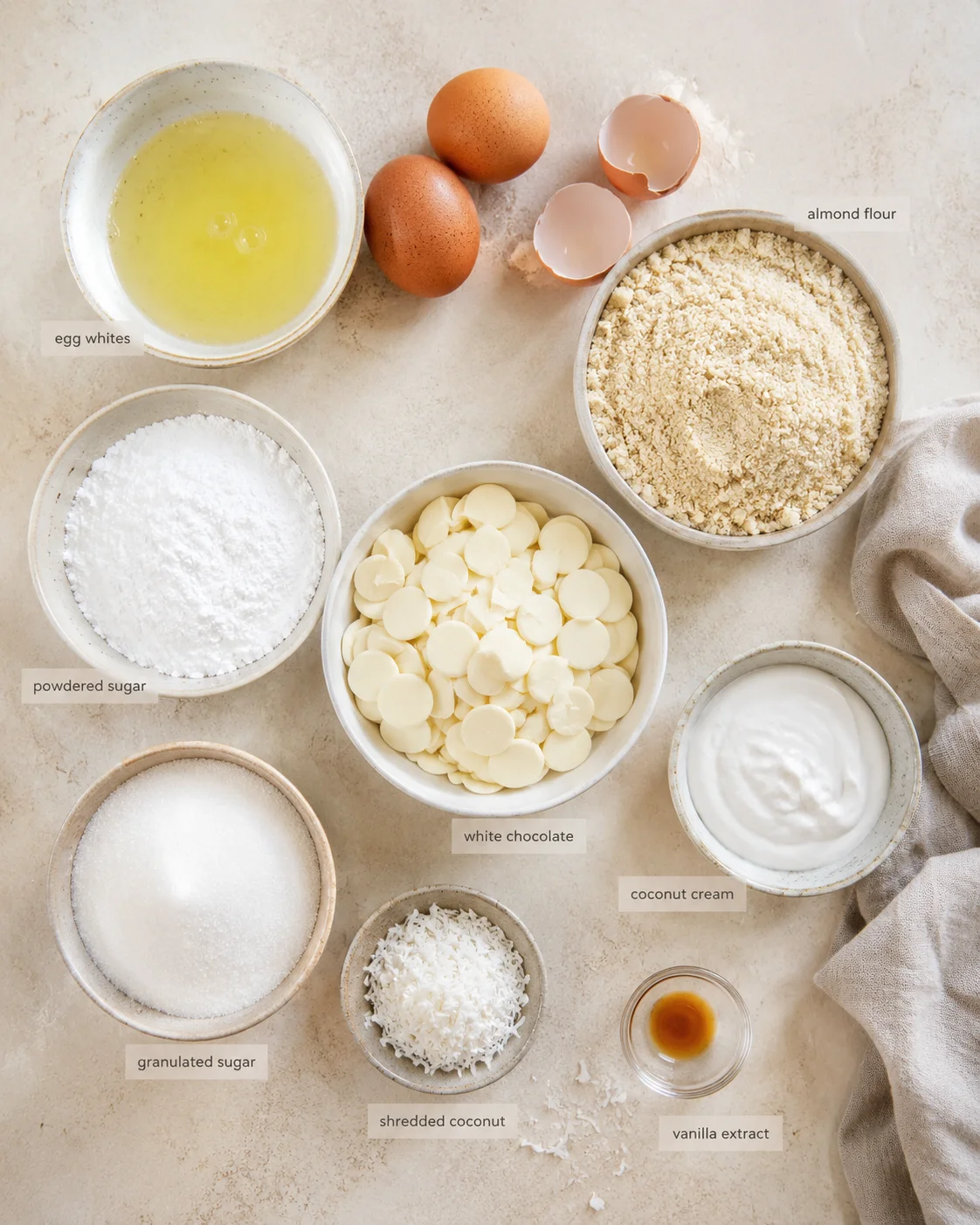 top-down flat lay of baking ingredients labeled for a recipe: almond flour, sugars, white chocolate, shredded coconut, and eggs