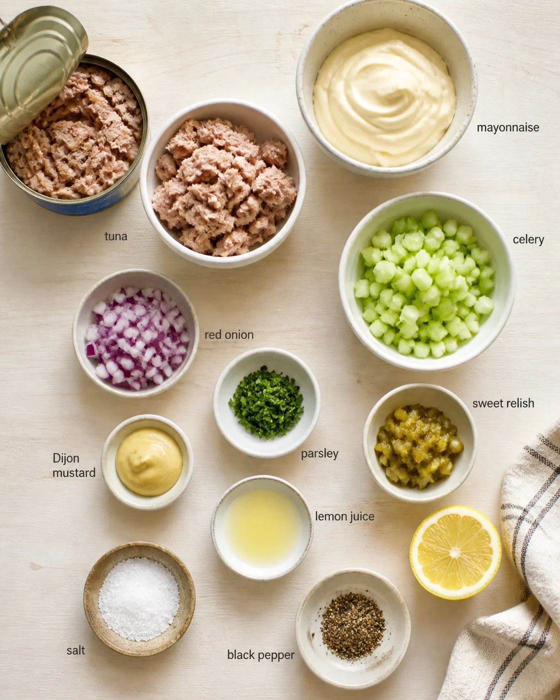 Overhead view of tuna salad ingredients arranged in bowls on a light wooden surface.