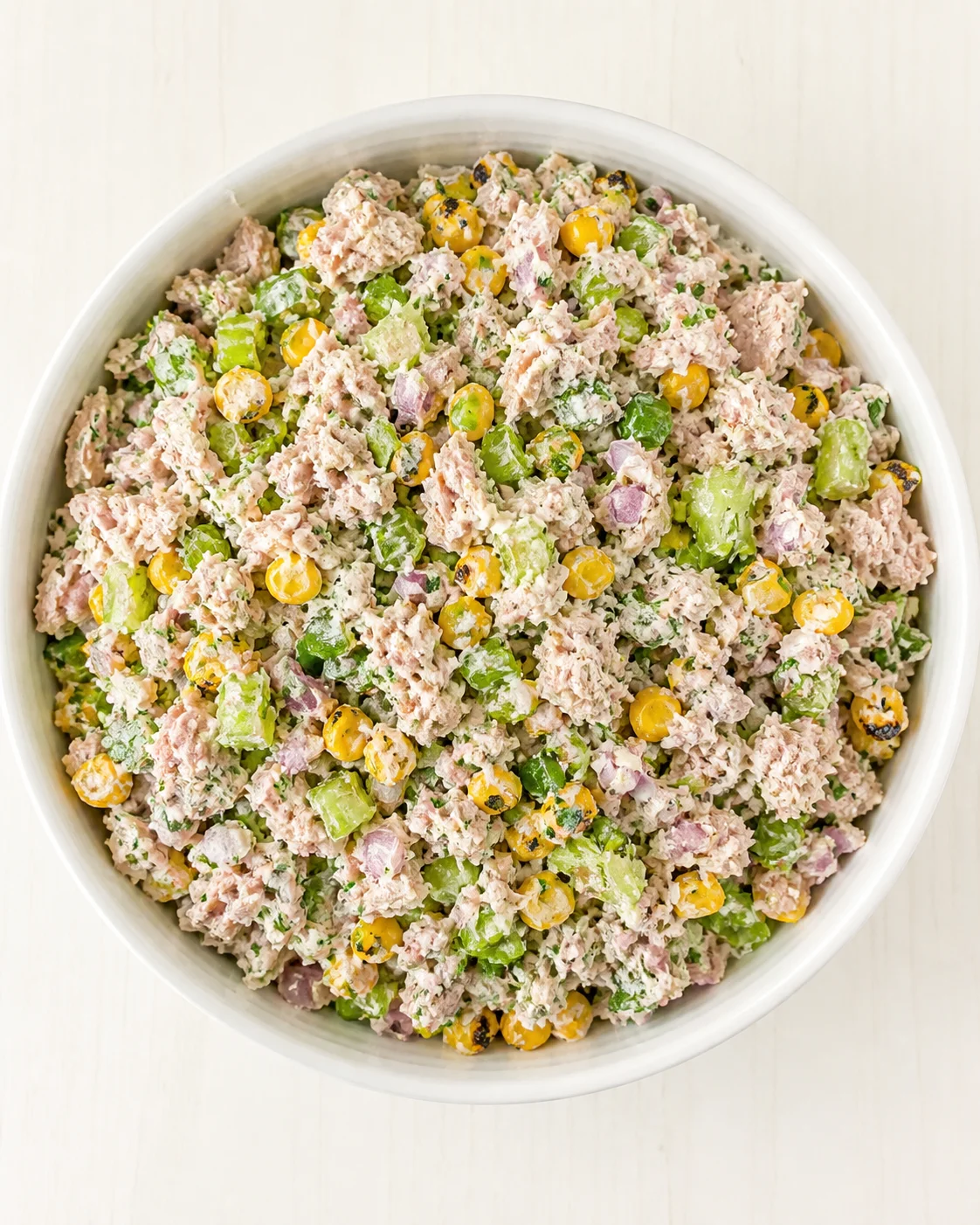 Top-down view of a white bowl filled with tuna salad with corn, celery, and red onion.