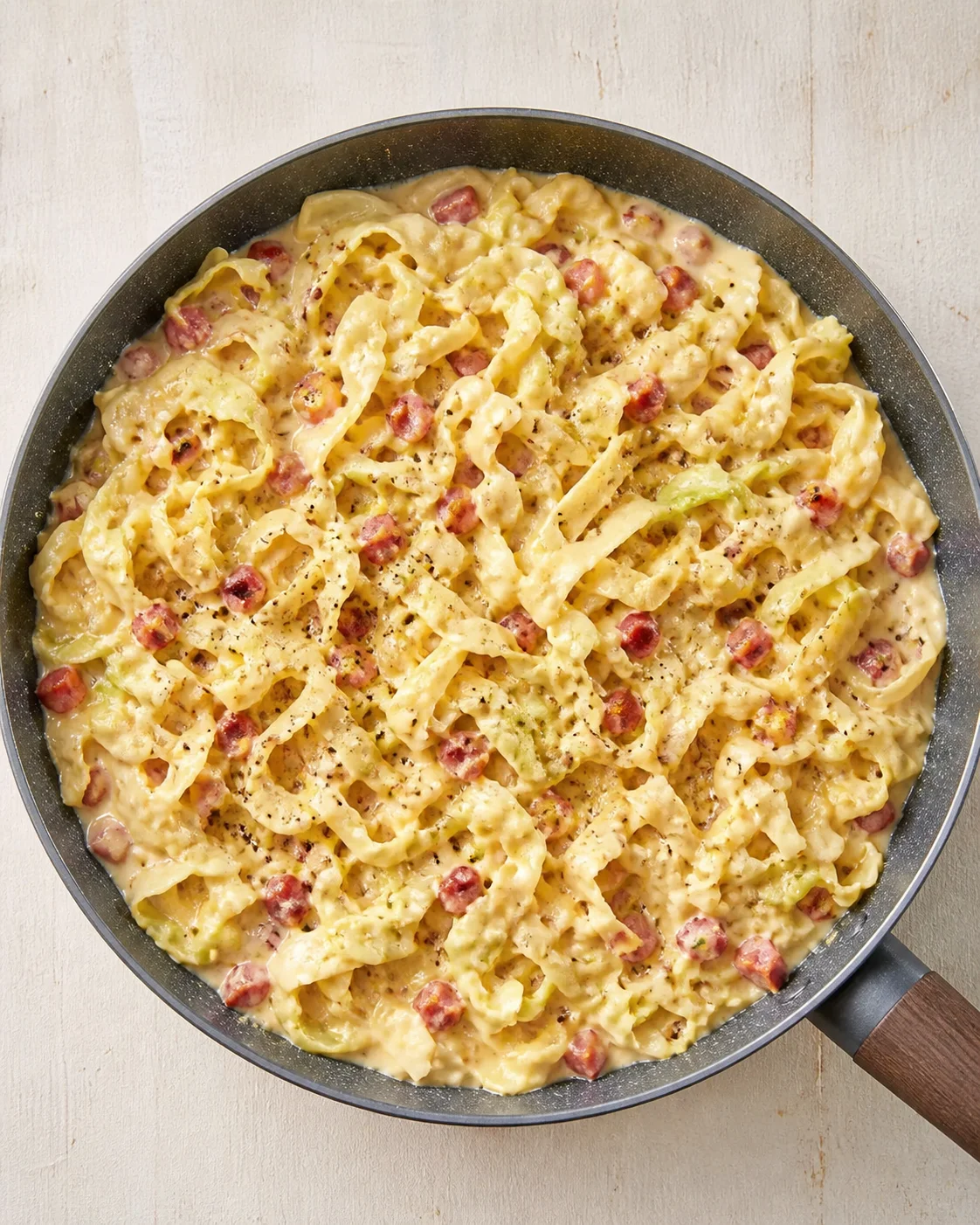 Top-down view of creamy sausage pasta with pink sausage chunks in a dark skillet, sprinkled with black pepper.