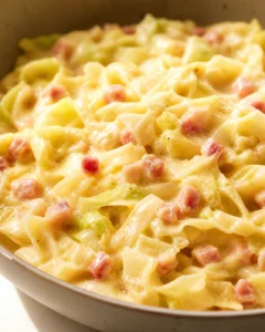 Close-up of creamy pasta with diced ham and pale-green chunks in a skillet.