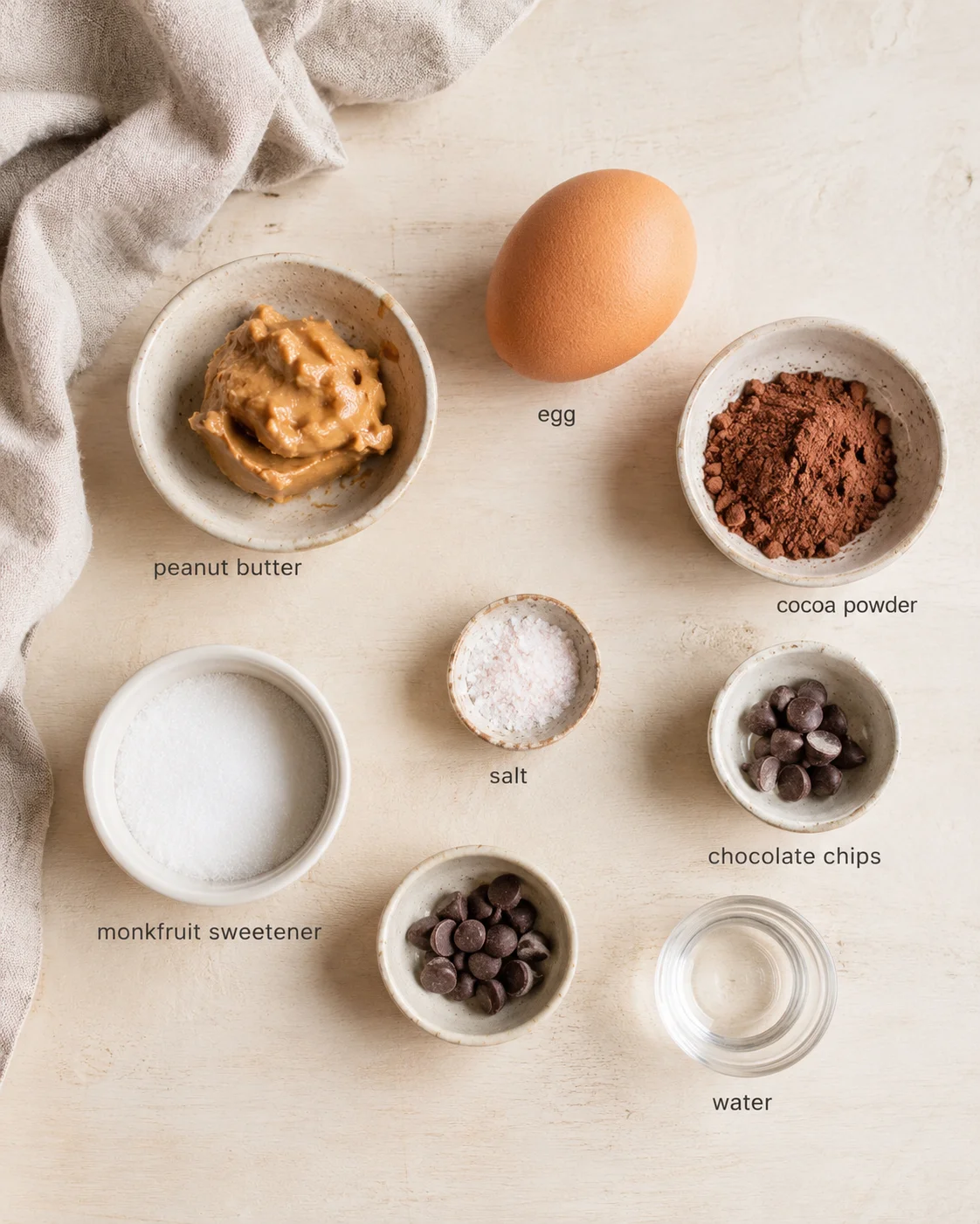 Flat lay of recipe ingredients on a light wooden surface, including peanut butter, egg, and cocoa powder.