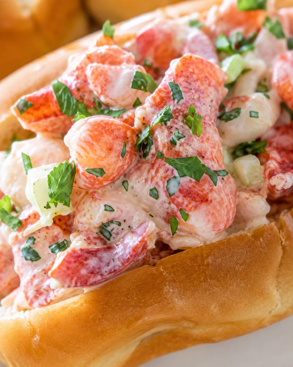 Close-up of a lobster roll with creamy mayo, chopped parsley, and diced celery in a toasted bun.