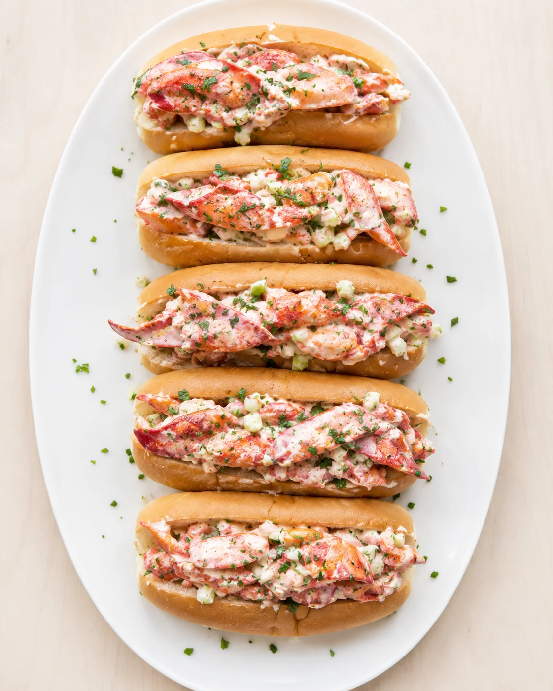Top-down view of five lobster rolls with creamy filling on a white oval plate.