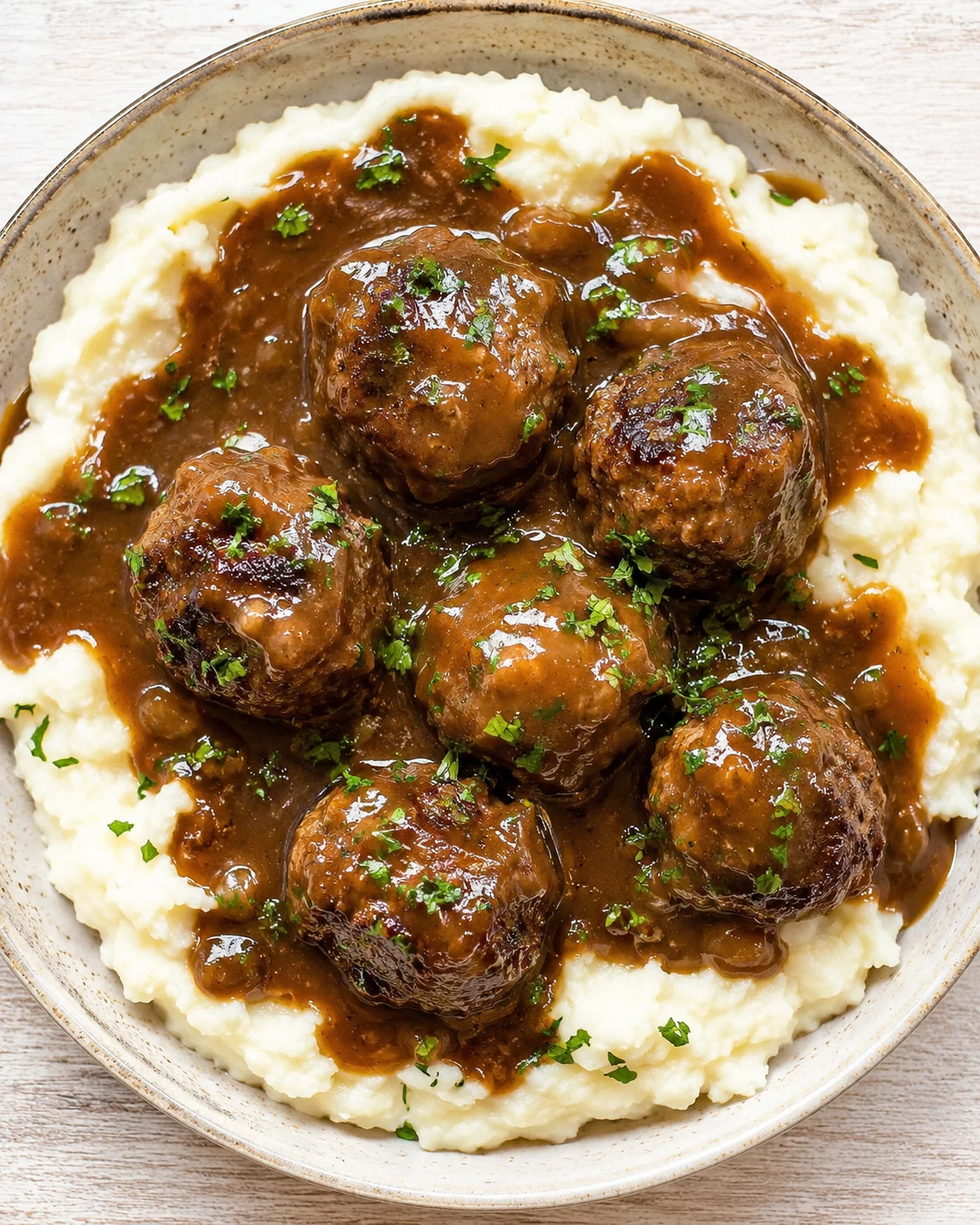 Creamy mashed potatoes topped with browned meatballs in brown gravy, garnished with parsley.