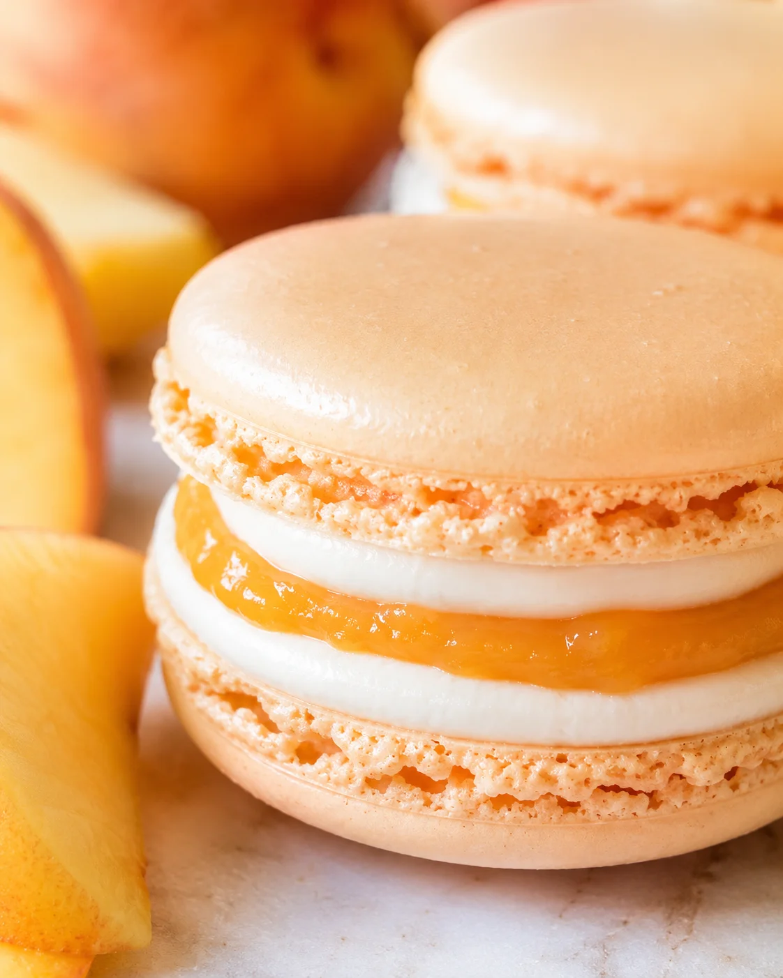 Close-up of peach macarons with white cream and orange jam filling, surrounded by peach slices.