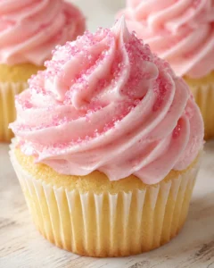 Close-up of a vanilla cupcake with swirled pink frosting and pink sugar crystals on top, shown on a light wooden surface with a soft background