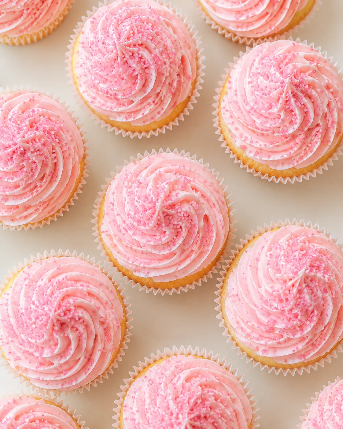 Close-up of pink cupcakes with swirled frosting and sugar sprinkles on a pale background.