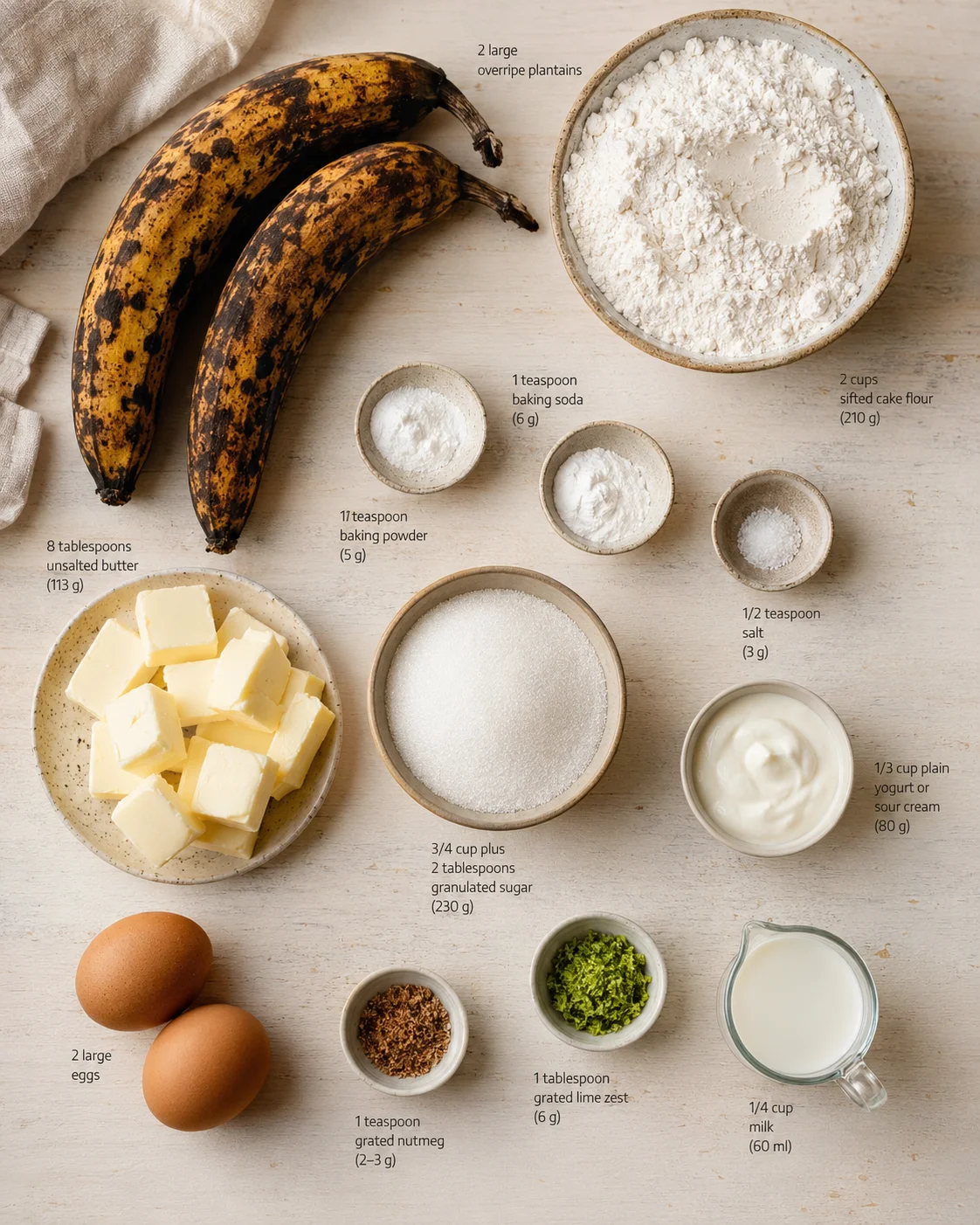 Overhead view of two overripe plantains surrounded by labeled bowls of flour, sugar, butter, eggs, yogurt, and milk.