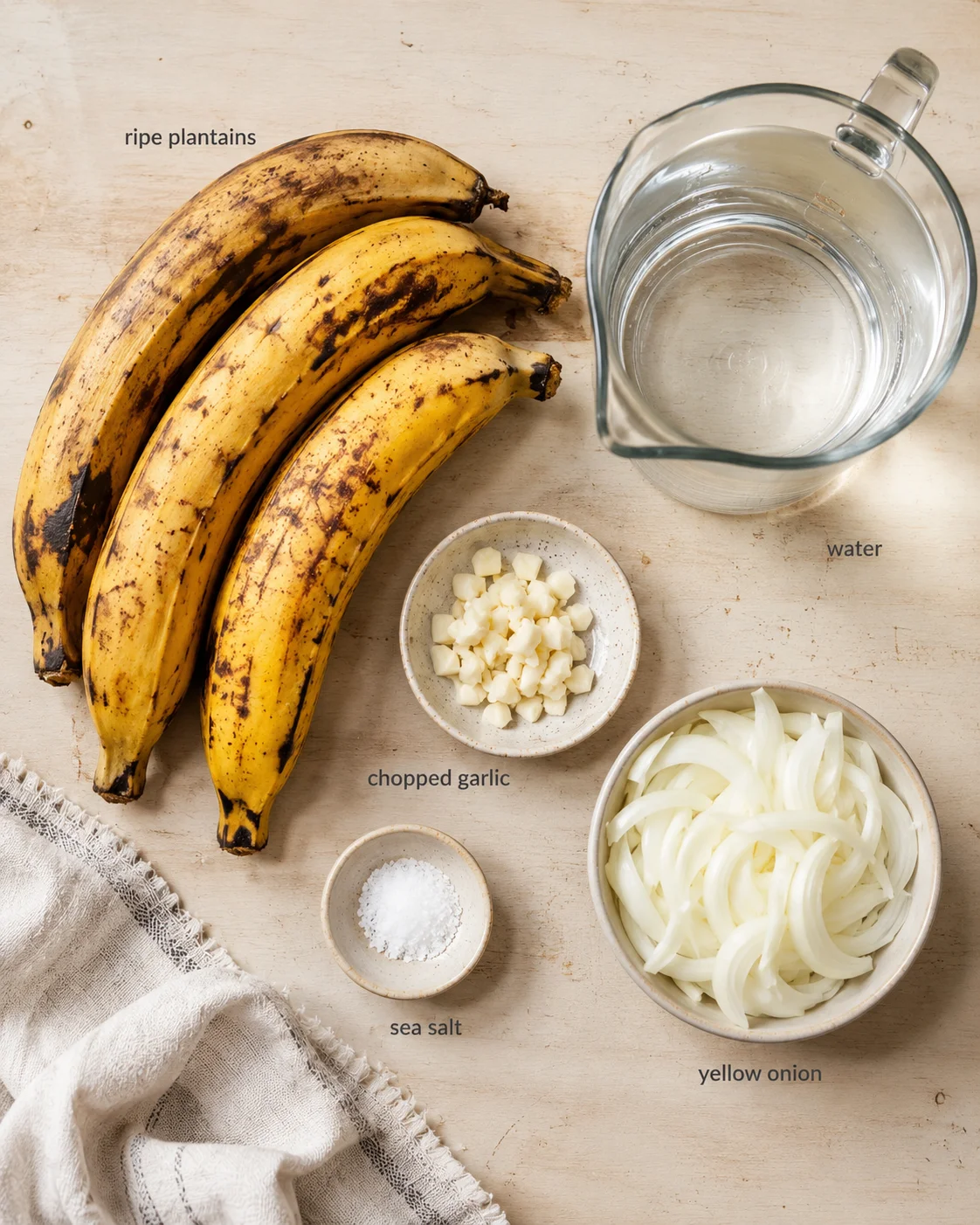 Overhead view of ripe plantains with water pitcher, chopped garlic, sea salt, and sliced yellow onion on a wooden surface.