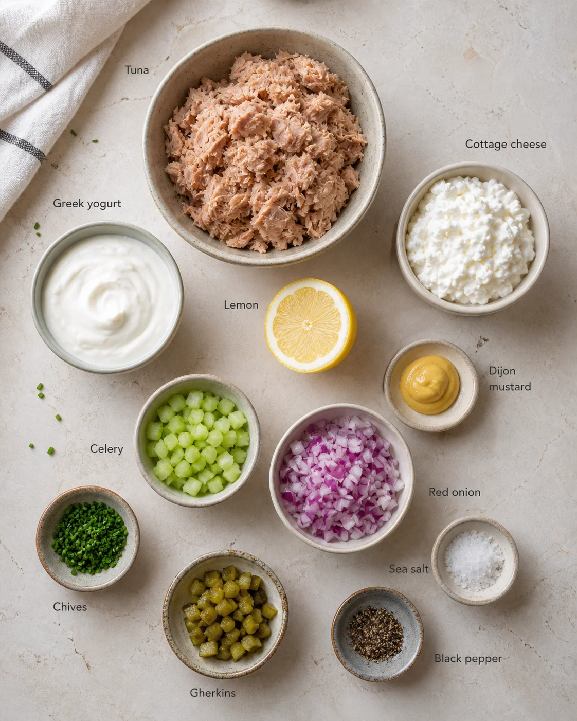 Overhead view of tuna, yogurt, cottage cheese, lemon, and chopped vegetables arranged on a pale stone surface.