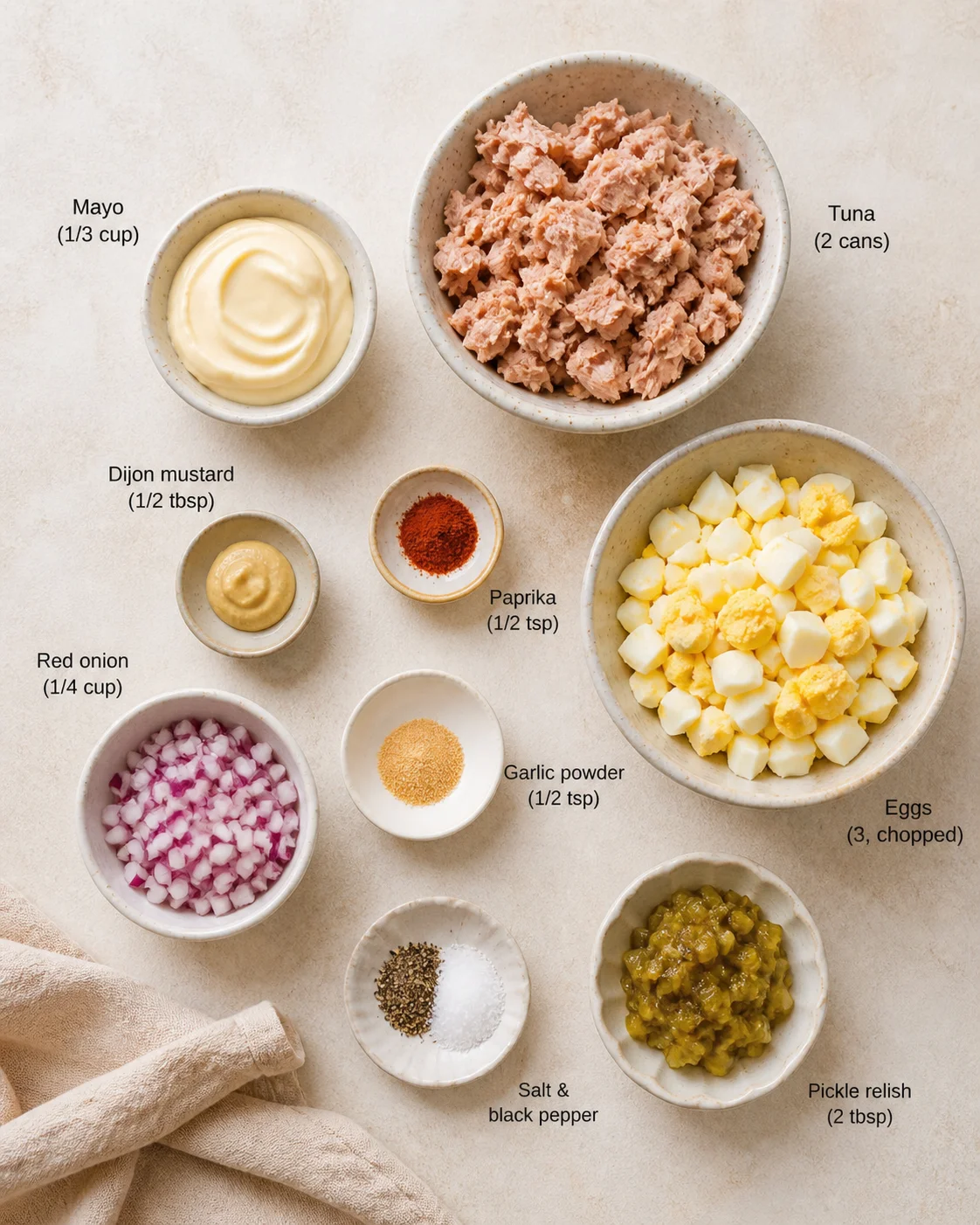 Top-down view of bowls with tuna, chopped eggs, mayo, red onions, and seasonings on a beige surface.