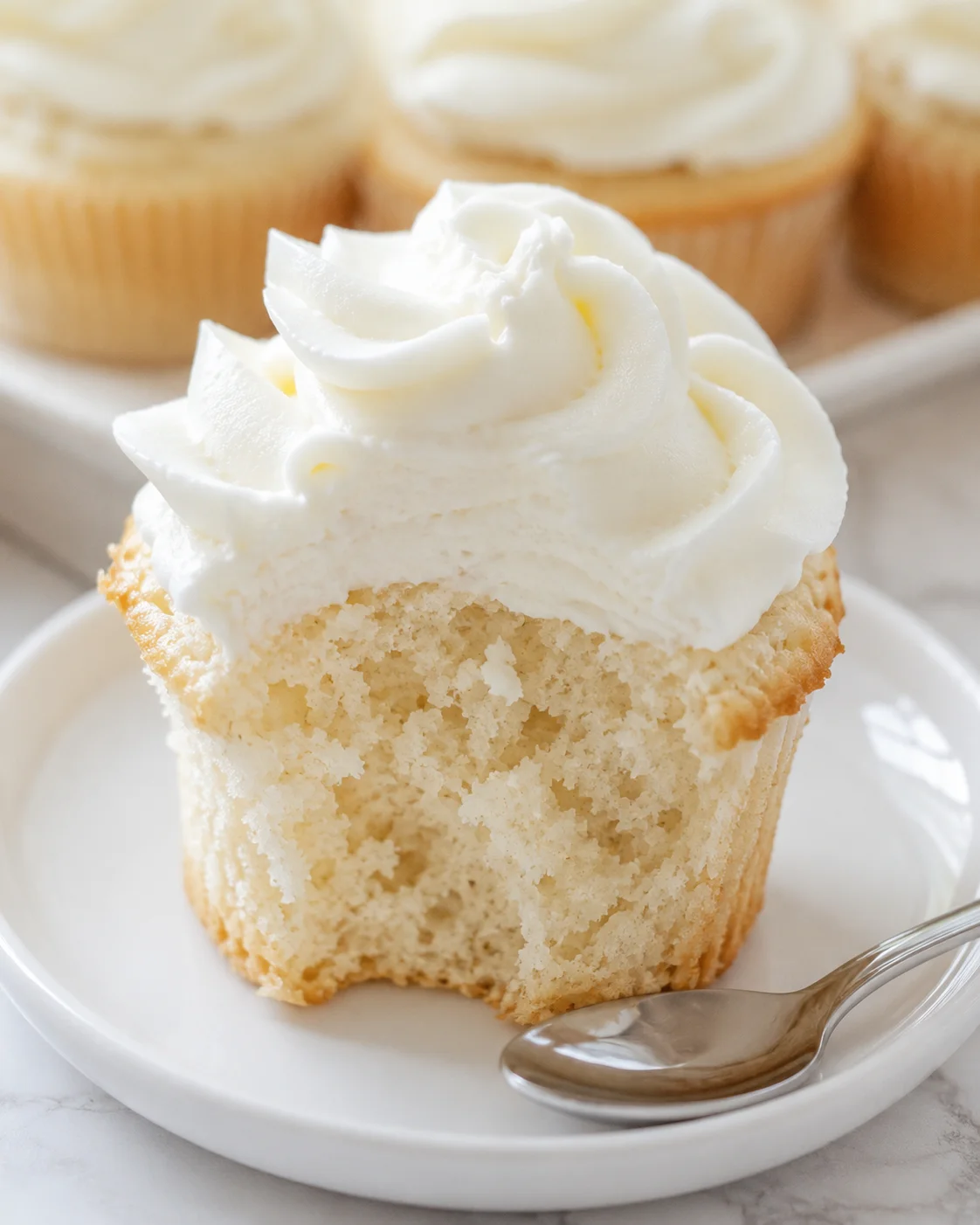 Close-up of a vanilla cupcake with white frosting on a white plate; airy crumb visible
